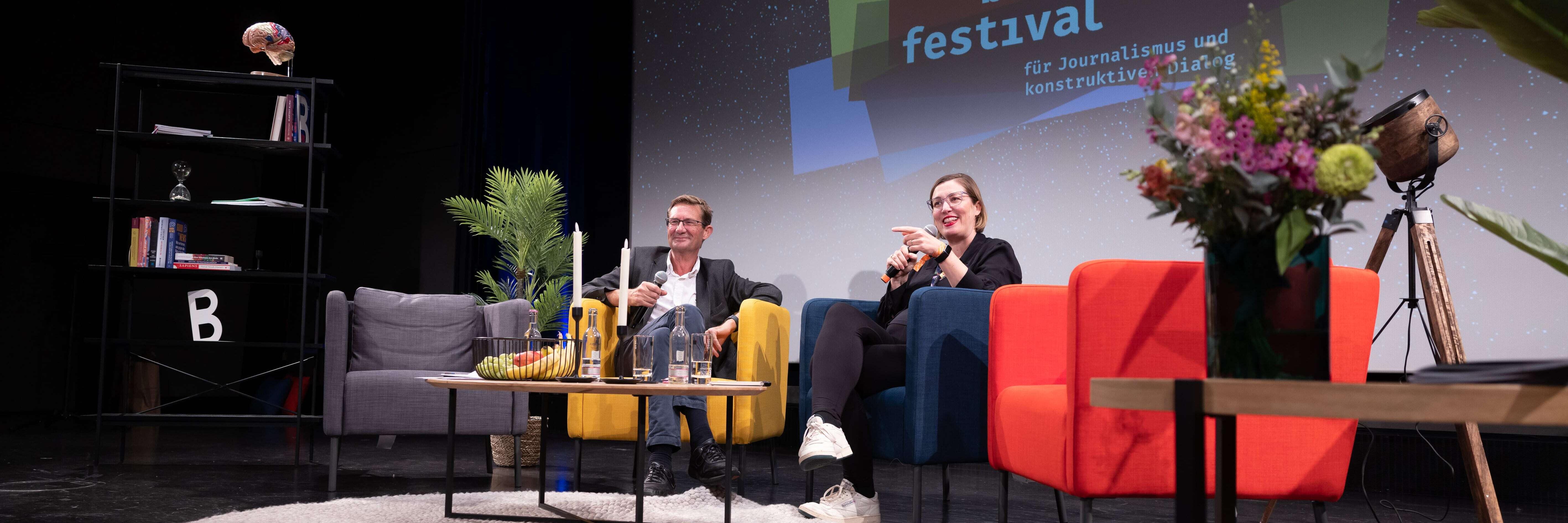 Nic Newman und Anita Zielina auf der Bühne im Gespräch, umgeben von bunten Requisiten.