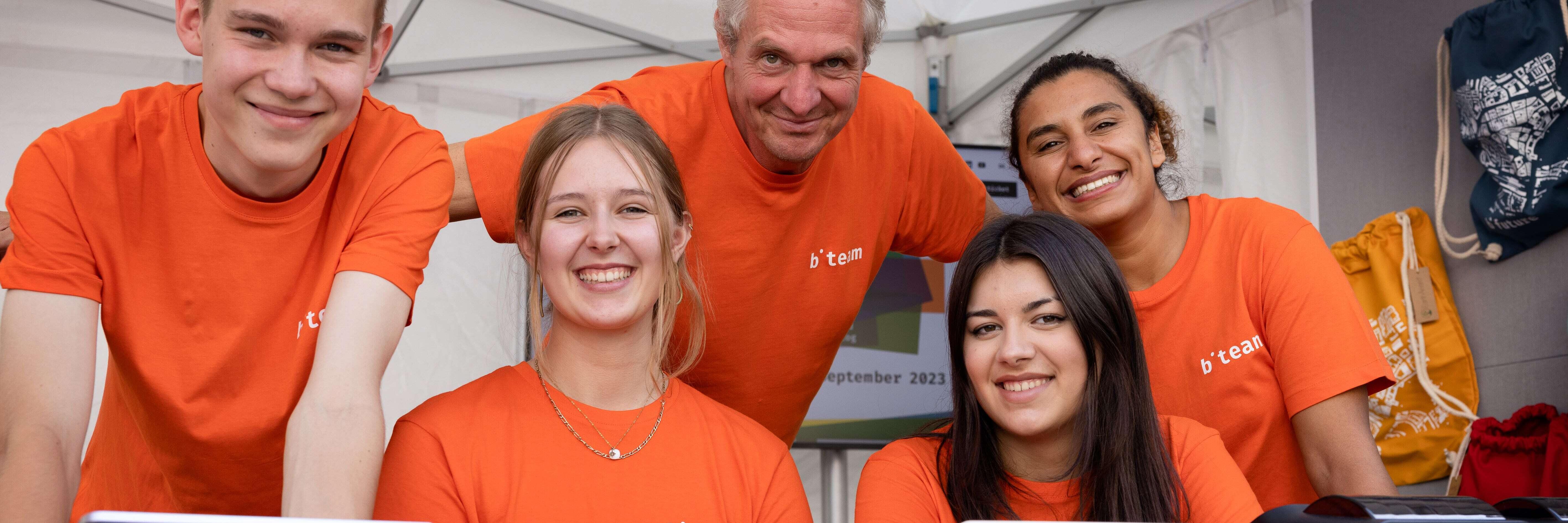 Five team members of the b-future team, dressed in orange, stand at a festival booth, smiling warmly at the camera. They are wearing orange T-shirts with the inscription "b-team."