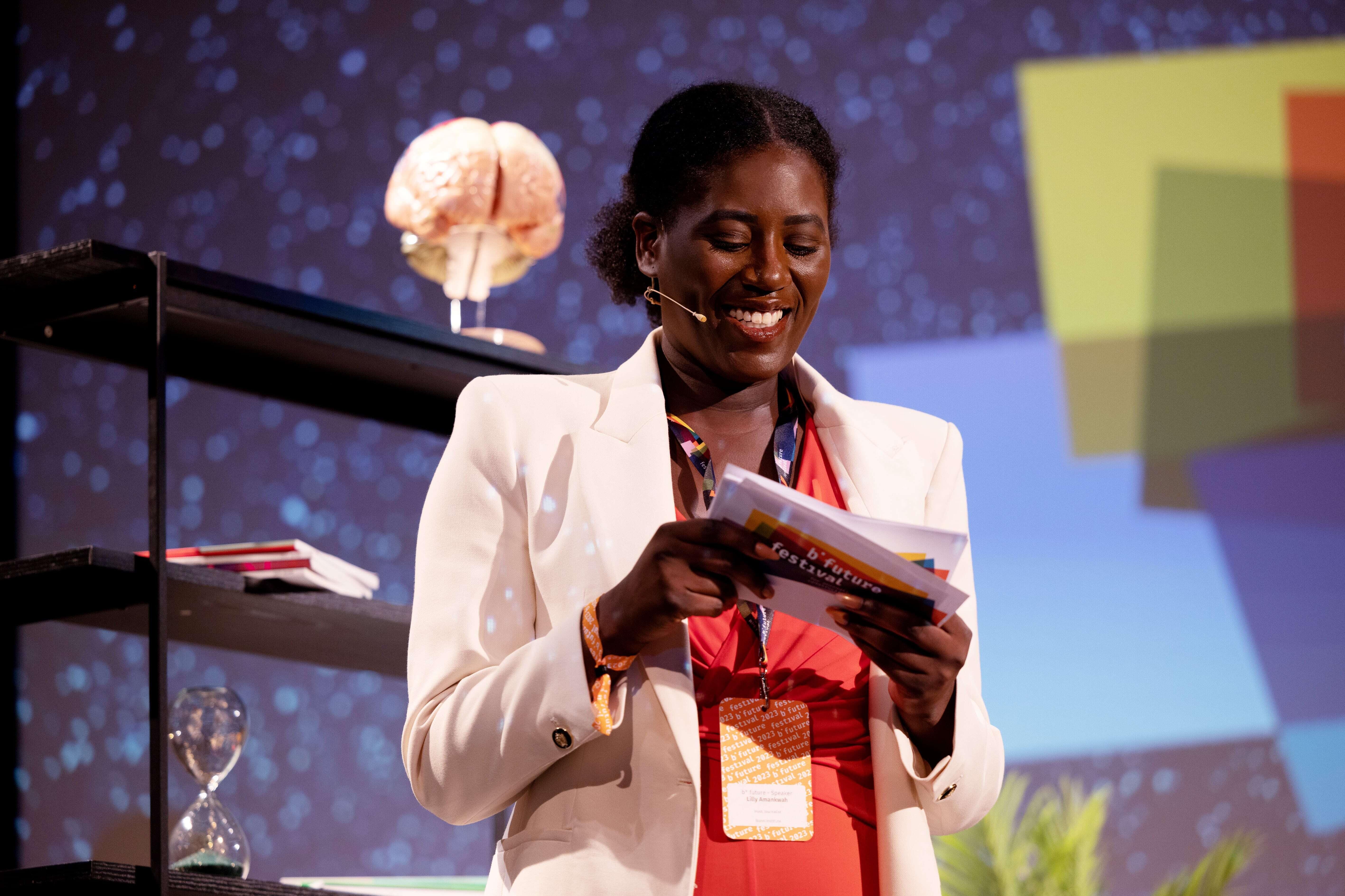 Lilly Amankwah steht auf der Bühne und hält Moderationskarten in der Hand, links von ihr ein Regal mit dem Modell eines Gehirns, im Hintergrund das b° future Logo auf einer Leinwand.