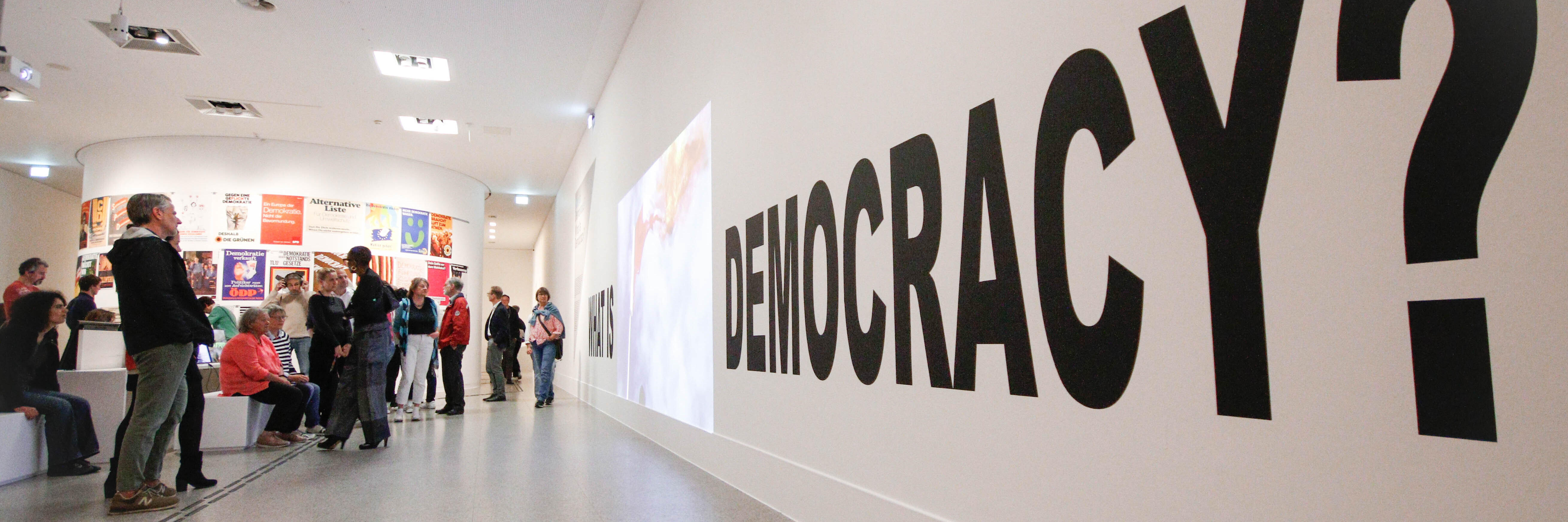 Ein Bild aus der Demokratieausstellung der Bundeskunsthalle. Zusehen sind Menschen die vor einer Wand stehen, an der groß Democracy steht.