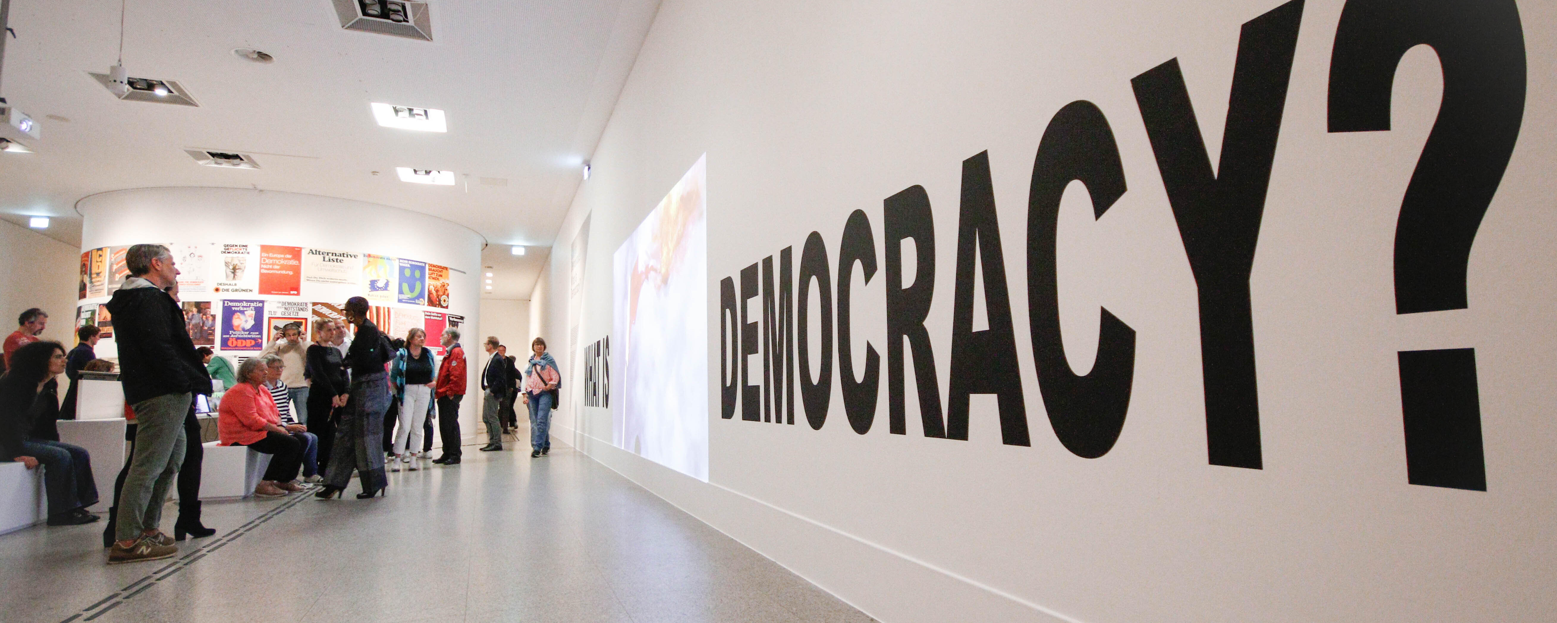 Ein Bild aus der Demokratieausstellung der Bundeskunsthalle. Zusehen sind Menschen die vor einer Wand stehen, an der groß Democracy steht.